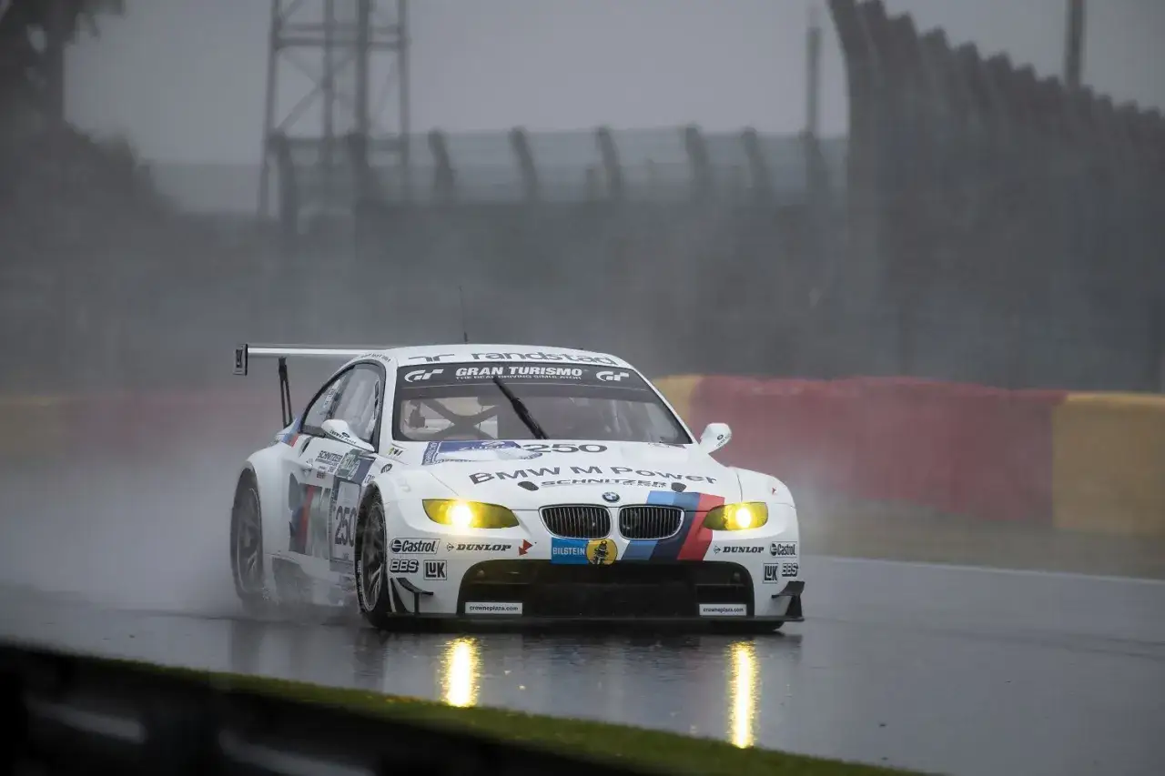 Ein weißer BMW M3 GT2 rast bei Regen über die nasse Rennstrecke, die Scheinwerfer leuchten hell.