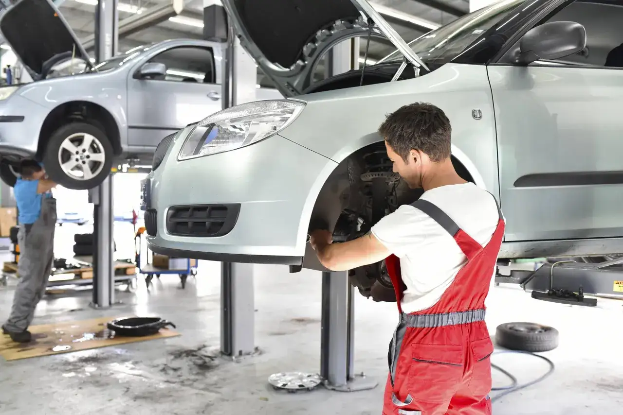 Mechaniker in roten Latzhosen wechselt an einem Auto die Bremsscheiben. Ein zweites Auto steht daneben auf einer Hebebühne.