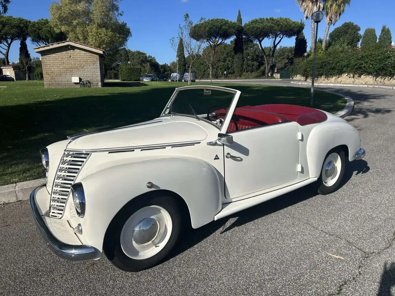 Ein weißer Biagini Passo Cabriolet mit rotem Verdeck steht auf einer Straße, umgeben von Bäumen und Grünflächen.