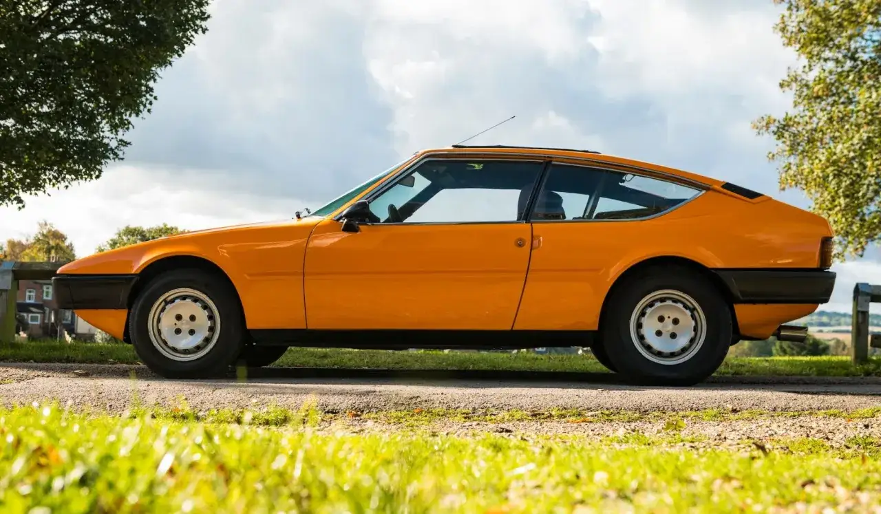 Orange Matra Bagheera Coupé parkt auf einer Straße, umgeben von Bäumen und Gras.