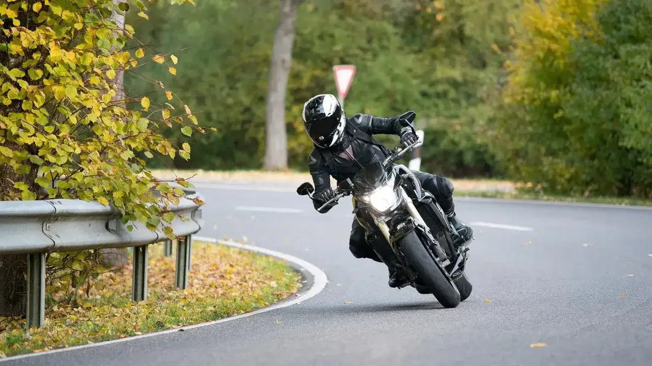 Motorradfahrer in voller Montur lehnt sich in einer Kurve, der Lenkimpuls des Motorrads ist deutlich zu sehen. Herbstlaub liegt am Straßenrand.