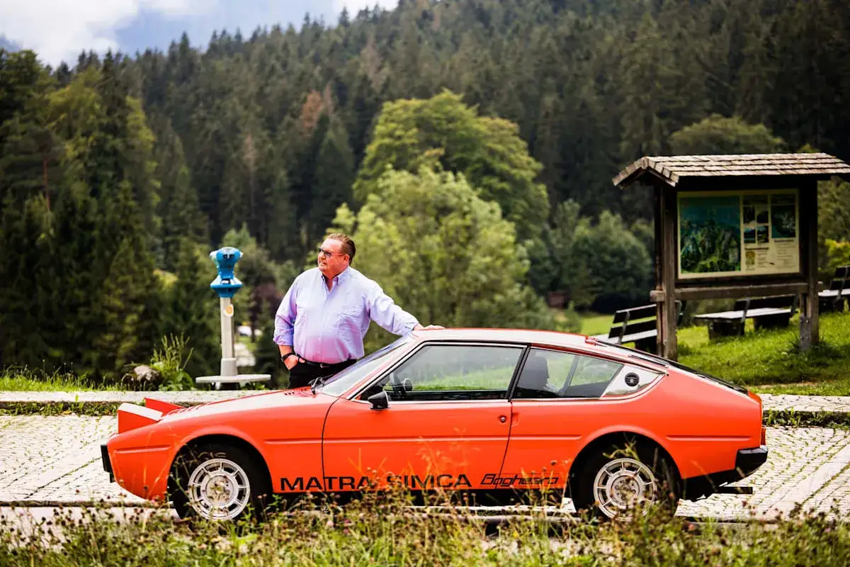 Mann lehnt an seinem knallroten Simca Matra Bagheera, umgeben von üppiger Natur und einem Bergpanorama.