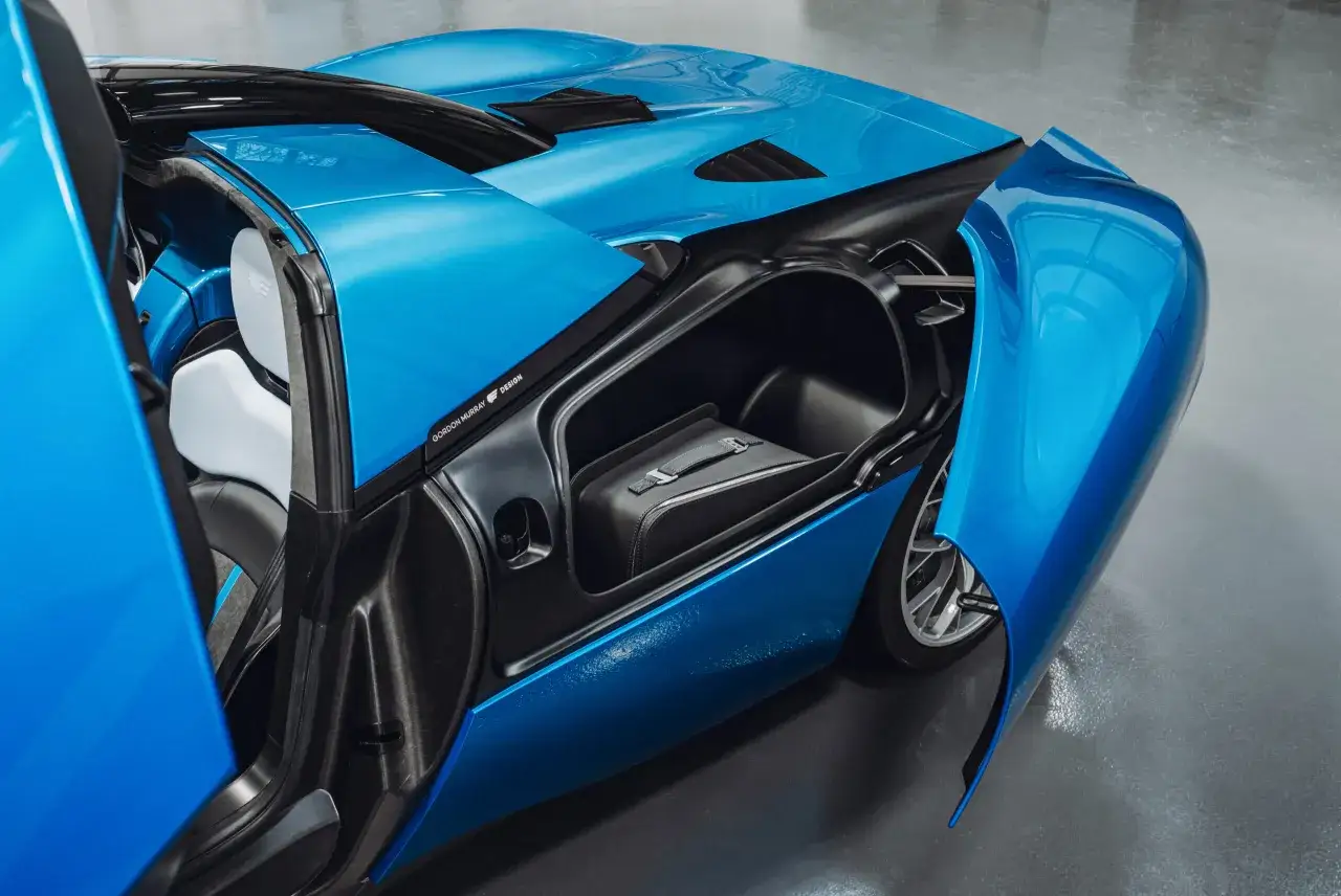 Der blaue Gordon Murray T.33 mit geöffneter Tür zeigt ein schwarzes Leder-Gepäckstück im Kofferraum.
