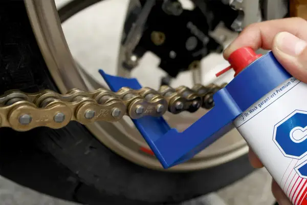 Hand sprüht mit einer Dose ein Motorrad Kette, um sie zu fetten. Ein blauer Kettenreiniger hilft dabei.
