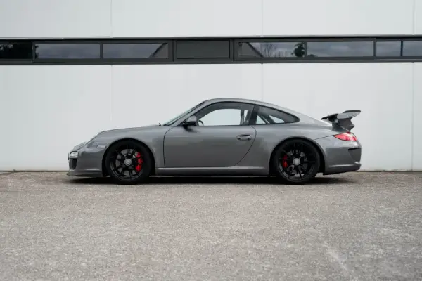 Grauer Porsche 997 GT3 RS mit schwarzem Flügel und roten Bremssätteln steht vor einer weißen Wand.